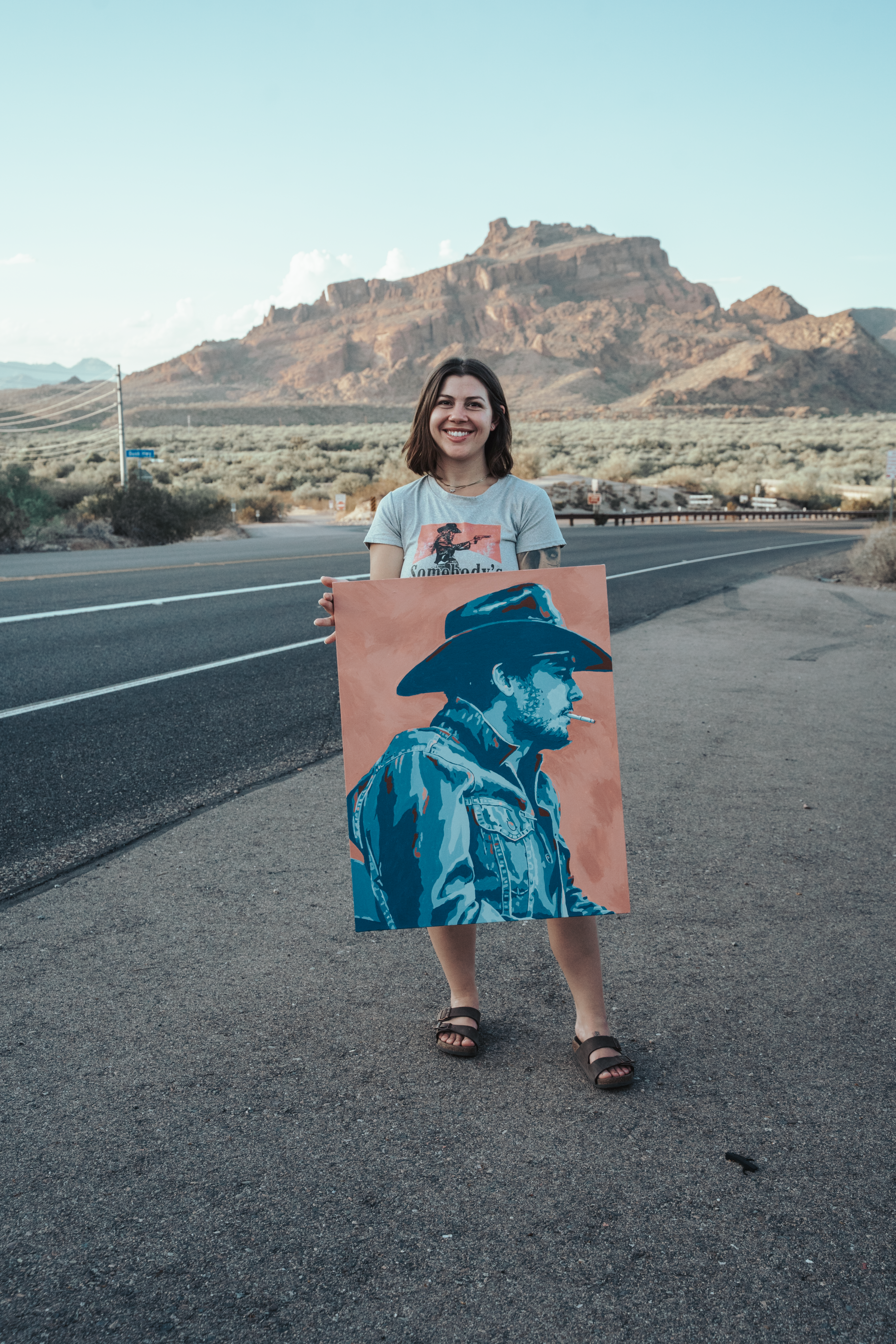 Jordan holding the cowboy painting on a desert highway, mountain in the distance