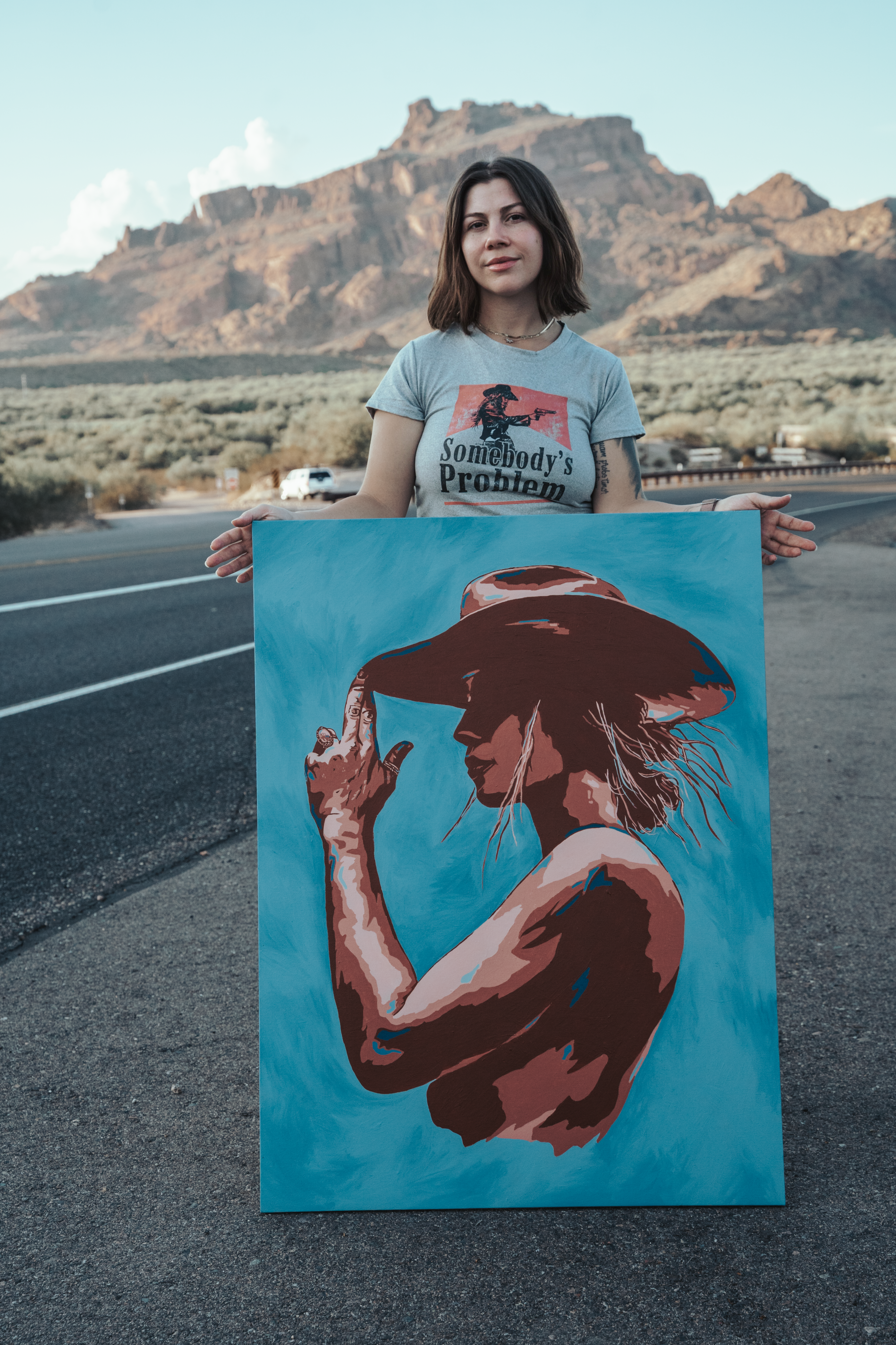 Jordan holding a Western portrait painting on a desert highway, mountain in the distance