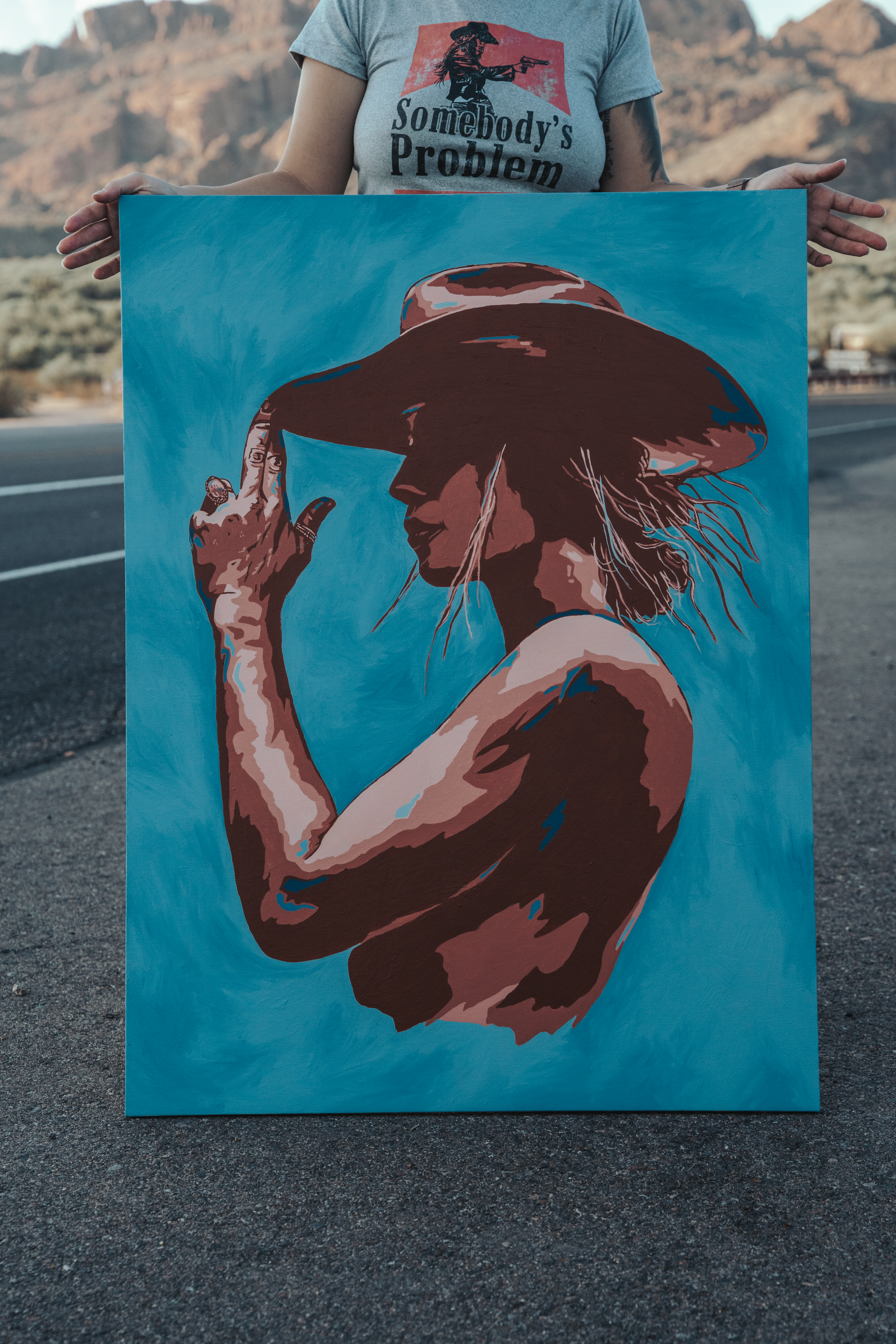 A large painted canvas held upright outdoors, hands visible at its edges to show scale