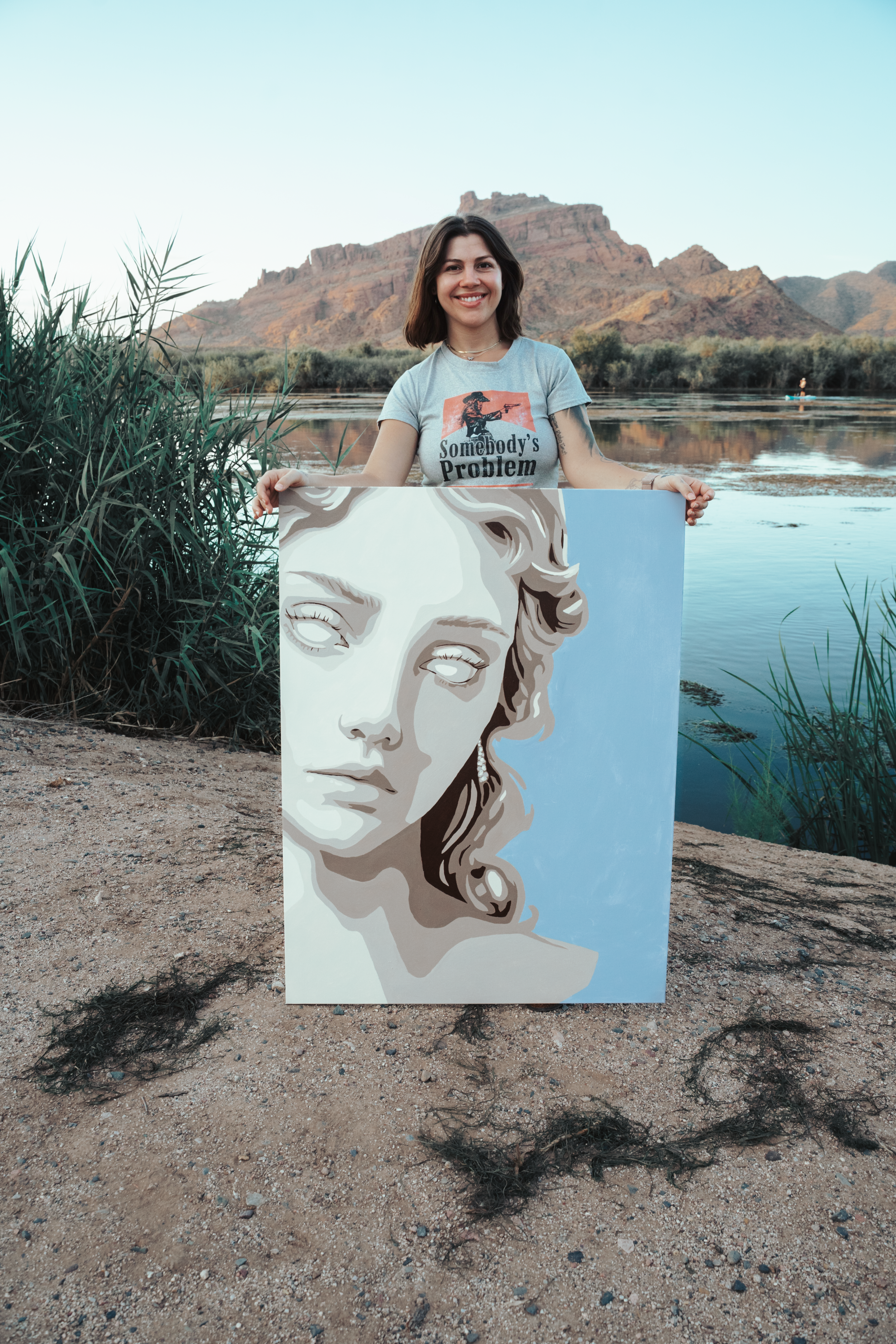 Jordan holding a classical portrait painting beside a desert river, reeds and mountain in the background