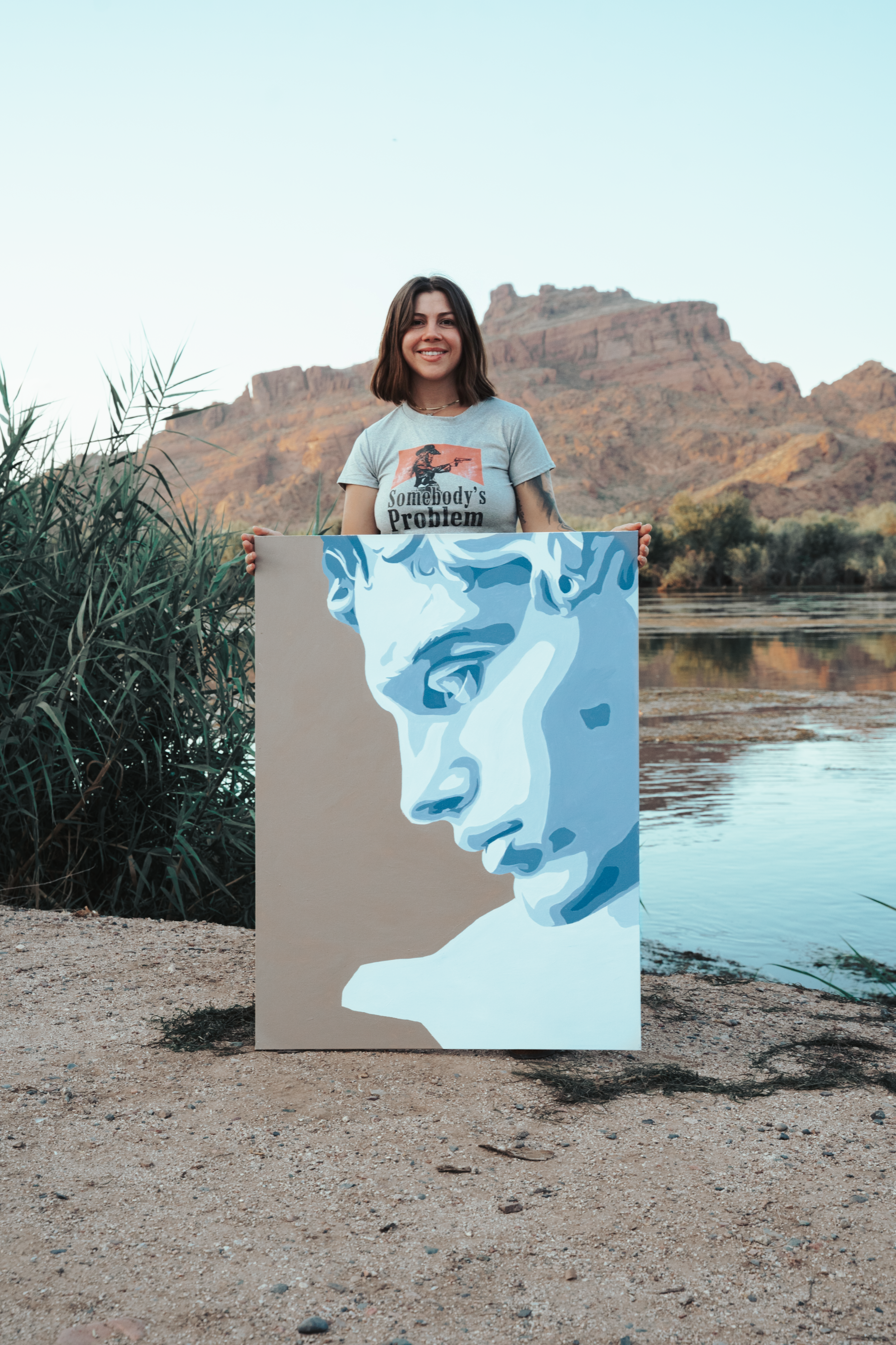 Jordan holding the classical portrait painting beside a desert river, reeds and mountain in the background