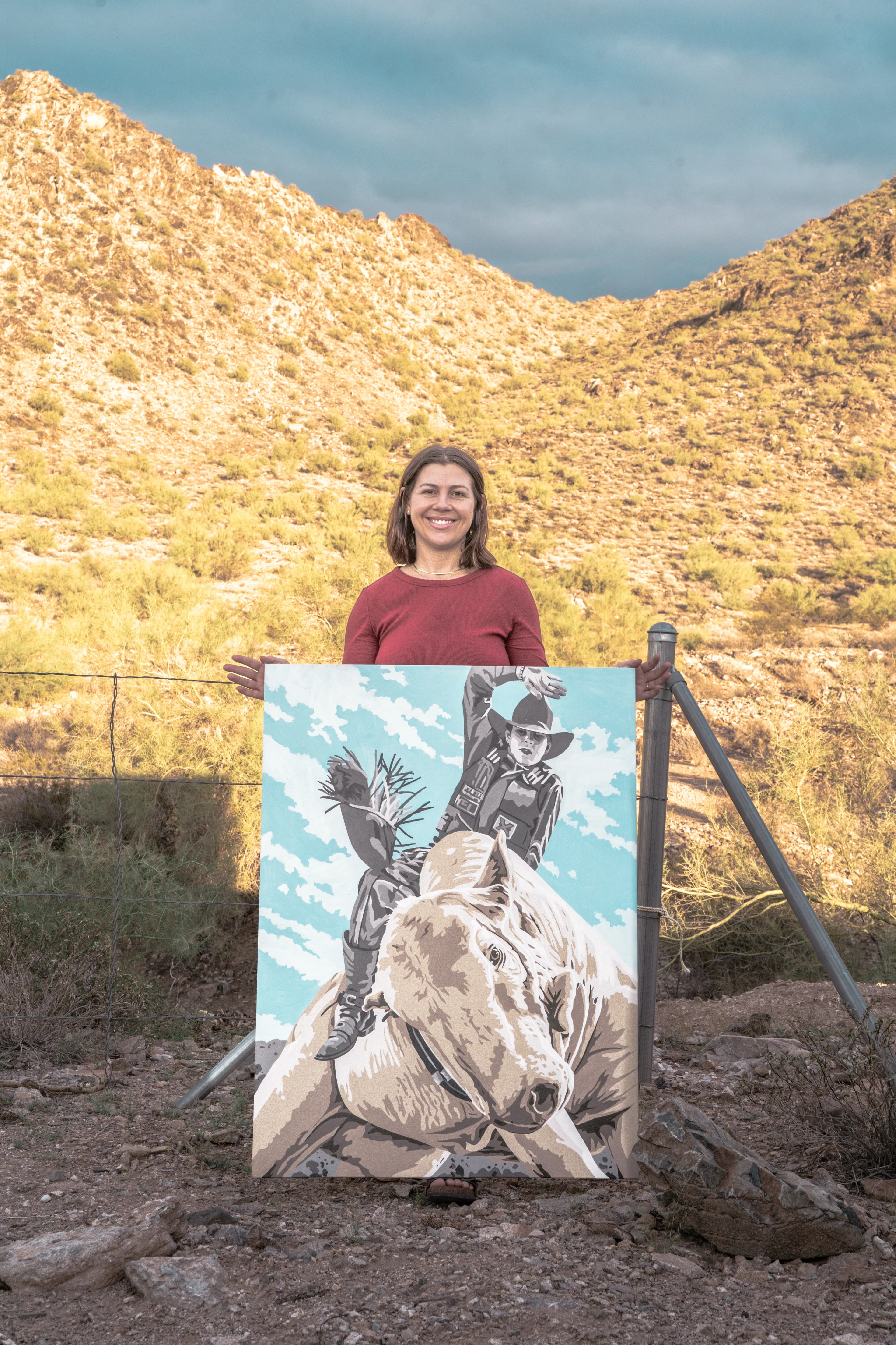 Holding the Buckin' Crimber painting at a wire fence, desert mountains in the background