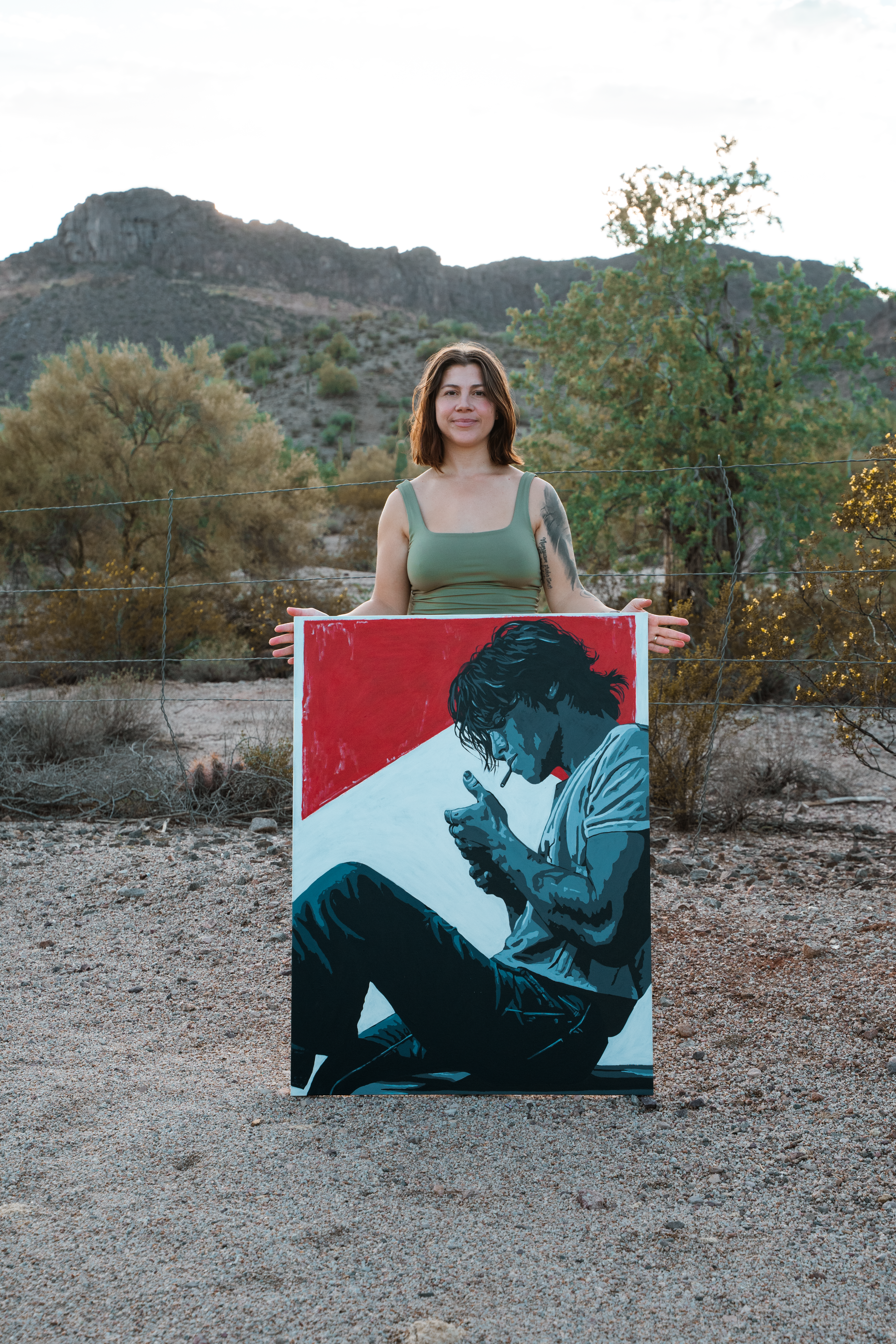Jordan holding the Nirvana painting at a wire fence, desert mountains in the background